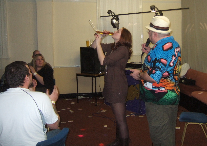 Contestant drinking from cock and ball shaped glass with Pav stood beside in loud shirt.